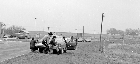 In this Bill Schock photo taken on Feb. 2, 1974, Kansas State Troopers crouch behind patrol cars during the shootout. Other police and patrol cars are strung out ahead. The overturned car in which the youths were fleeing is at the far upper right.